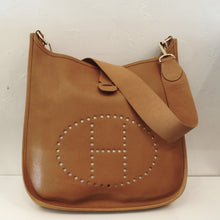 Load image into Gallery viewer, This Hermes Almond Evelyne Clemence Leather MM has gold hardware and a thick canvas shoulder strap. The top of the bag has a small strap pulling the top together and the back pocket has a small strap snaping it together. The interior of the bag is the underside of the leather and the outer side has a soft pebbly appearance.