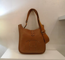 Load image into Gallery viewer, This Hermes Vintage Evelyne PM Clemence Leather 111-29 is in a deep camel color with silver hardware. The shoulder strap is adjustable and it has a thin strap attached to the back that goes through a front loop to secure the top of the bag. It has the hermes logo perforated on the front of the bag and a pocket on the back of the bag.