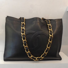 Load image into Gallery viewer, This Chanel Vintage Black Lambskin Leather Tote has the large Chanel Logo in raised letters on the front of the bag. It has gold hardware which includes the thick gold shoulder chain with a woven piece of leather through the chain links. The interior of this bag is also black.
