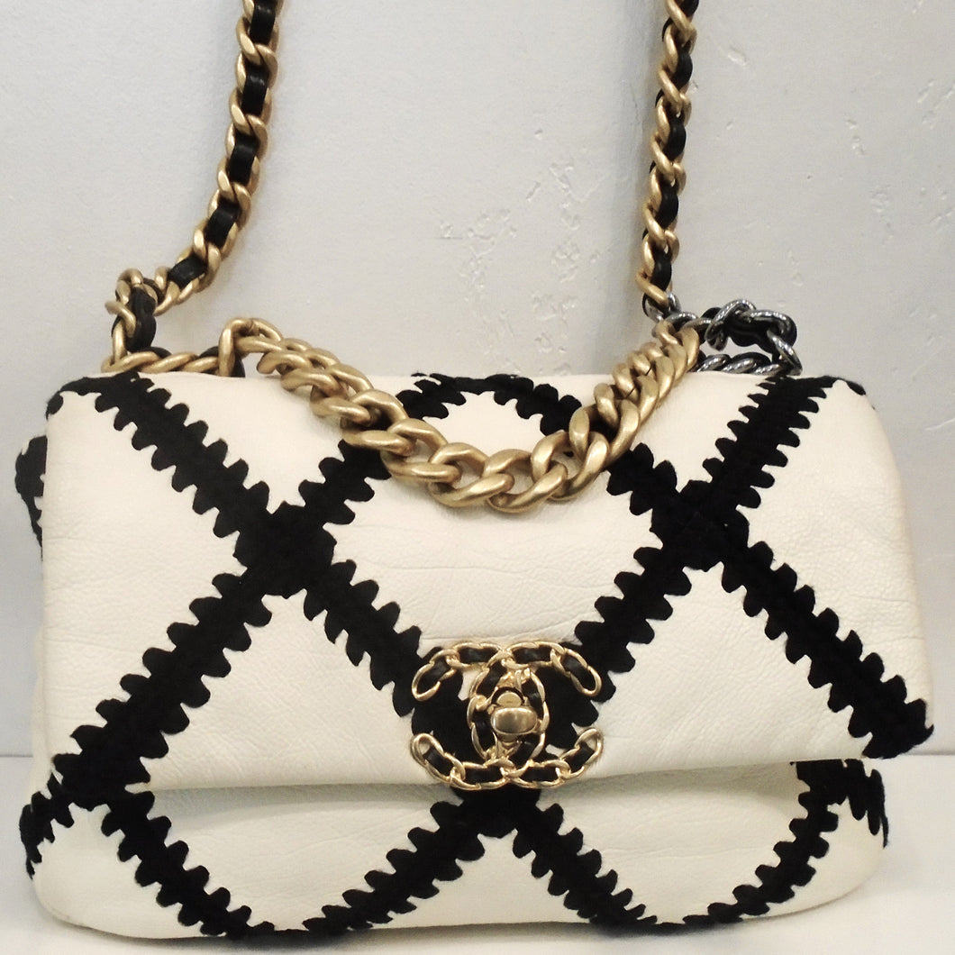 This Chanel Vintage  19  Two Tone Cotton & Leather Bag has black strips of fabric with chevron edges in the chevron design on the bag. The bag is white and has three tone gold hardware. It has the double C chanel logo in gold on the front center of the bag. There is a pocket on the outside back of the bag and the interior is white.