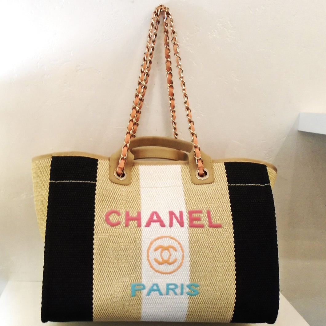 This Chanel Vintage Deauville Straw Tote is a woven bag with black, gold and white color strips with the white being the center. Chanel Paris and the CC logo are stitched on the front of the bag . Chanel stitched in pink, the CC logo in gold and Paris stitched in sky blue. It has a gold shoulder chain with a peach leather strip woven between the links. This bag also has two carrying handles. The interior of this bag is a soft pink.