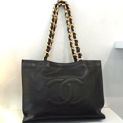 This Chanel Vintage Lambskin Large Tote has gold hardware (gold chain carrying handles with a black leather strip woven between the links). It has the Chanel CC logo raised on the front center of the bag. 