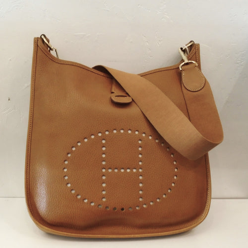 This Hermes Almond Evelyne Clemence Leather MM has gold hardware and a thick canvas shoulder strap. The top of the bag has a small strap pulling the top together and the back pocket has a small strap snaping it together. The interior of the bag is the underside of the leather and the outer side has a soft pebbly appearance.