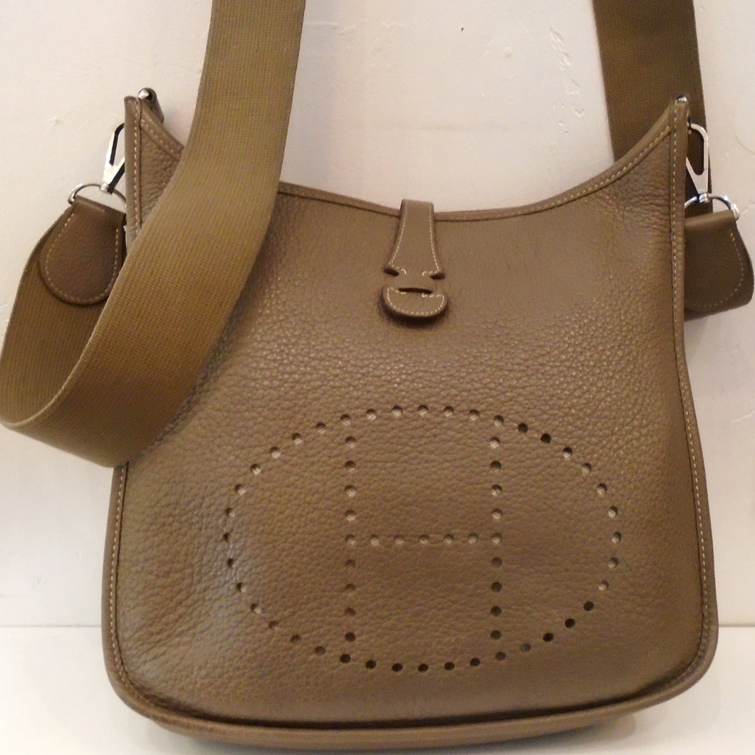 This Hermes Vintage Etoupe Evelyne PM Clemence Leather has silver hardware and the preforated Hermes Logo on the front of the bag. It also has the canvas (guitar strap) shoulder strap.