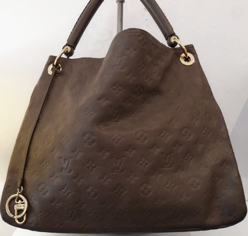 This Louis Vuitton Vintage Brown Empreinte Leather Artsy MM has a heavy duty leather carrying handle, pleated sides and a key ring attached to a leather strip on the outside of the bag. It has a cloth interior that is brown with black stripes.
