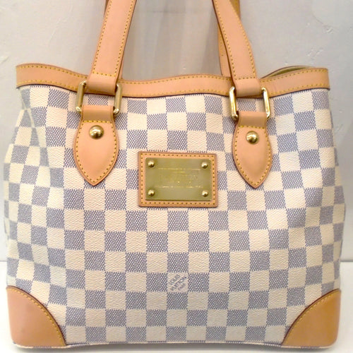This Louis Vuitton Vintage Damier Azur Hampstead PM has camel trim and gold hardware. It has leather on the outside corners of the bag and double carrying handles. The outside top corners have snaps to help close the top opening. It has a gold Louis Vuitton Plaque on the front of the bag and the interior is a soft cream color with inside pockets.
