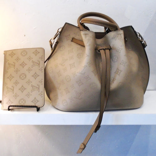 This Louis Vuitton Vintage Mahina Girolata With Pochette is a large drawstring bag with dark camel leather strips knotted at the ends for the drawstring. It has double handles, also in the dark camel leather. The bag is a light brown background with monogram print in a little darker brown.  The interior of the bag is a grey-brown with monogram pattern and Louis Vuitton printed on the bottom of the bag.