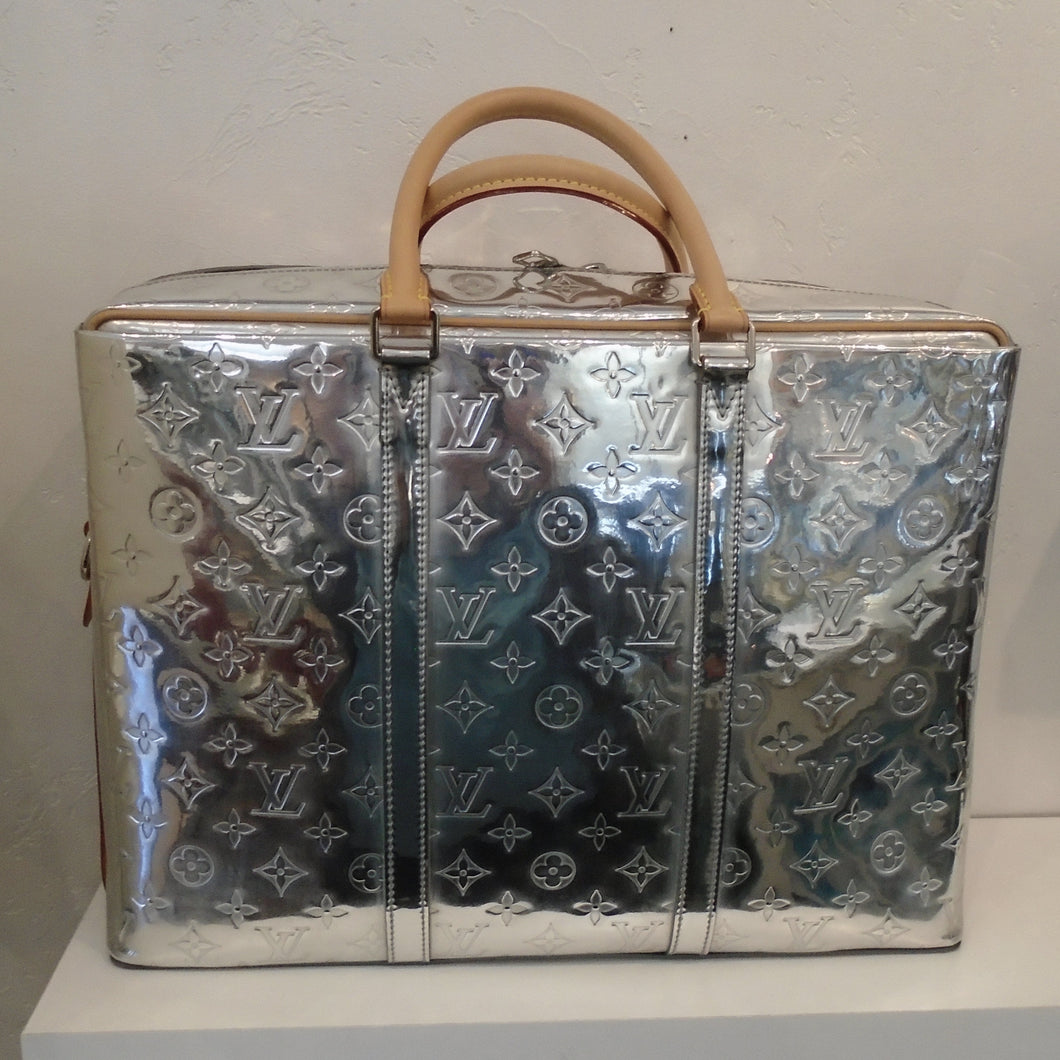 This Louis Vuitton Vintage Mirror Porte Documan Voyage Brief Case is a shiny silver with small Louis Vuitton monogram imprinted in the body of the bag. It has two carrying handles which are camel colored and includes a camel shoulder strap. The interior of the bag is a soft silver.