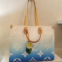 Load image into Gallery viewer, This Louis Vuitton Vintage On The Go GM has large white monogram on a blue faded into pink bag with camel leather trim. It has a daisy hanging from a leather strip from the front carrying handle. This bag has two carrying handles and two shoulder straps. The interior of the bag is light pink with white stripes.
