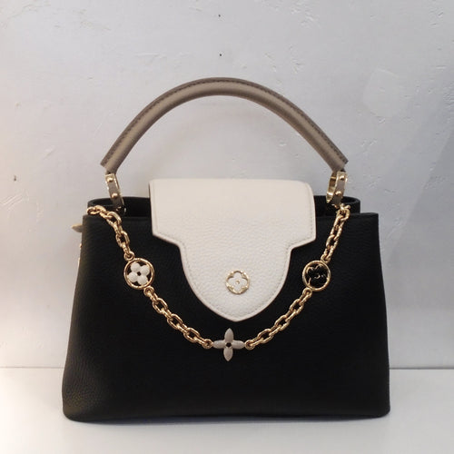 This Louis Vuitton Tricolor Taurillon Capucines MM is Black with a white flap that has a lip with a flower outlined in gold toned hardware on the lower lip. A chain drapes just under the lip with three flowers which is also gold toned hardware. It comes with a gray carrying handle. The interior of this bag is also black with sections one of which is zippered.