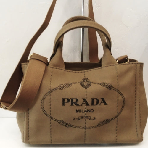 This Prada Vintage Canvas Canapa Handbag with Strap is a light shade of camel with dark brown print. The front of the bag has the Prada name, Milano and the date it was established on the front of the bag. It has a detachable sturdy canvas shoulder strap included and has gold hardware.