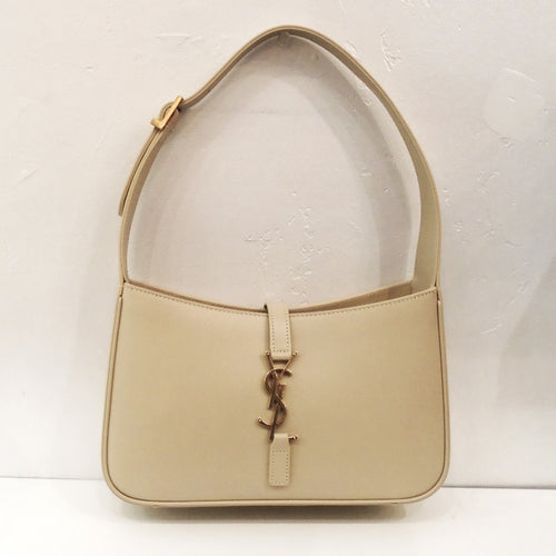 This YSL Cream Hobo Bag is a dark cream color with a thick adjustable shoulder strap. The top opening of the bag closes with a leather strip attached from the back to the front by a YSL logo hook into a bottom leather piece. The interior of the bag is the leather underside of the bag.