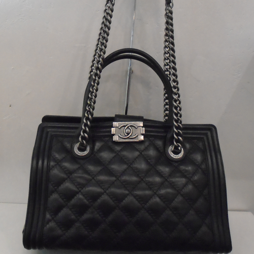 This Chanel Vintage Boi Tote has greyish silver hardware which includes the double shoulder straps with black leather handles at the top. It has black leather double handles for carrying by hand. It also has a black strap that secures the bag together in the top center of the bag with silver interlocking Cs on the buckle.