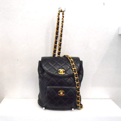 This Chanel Vintage Lambskin Backpack is Black with gold latches. It has a front pocket on the lower body of the bookbag. The straps are made of gold plated chain with a black leather strip woven through it.