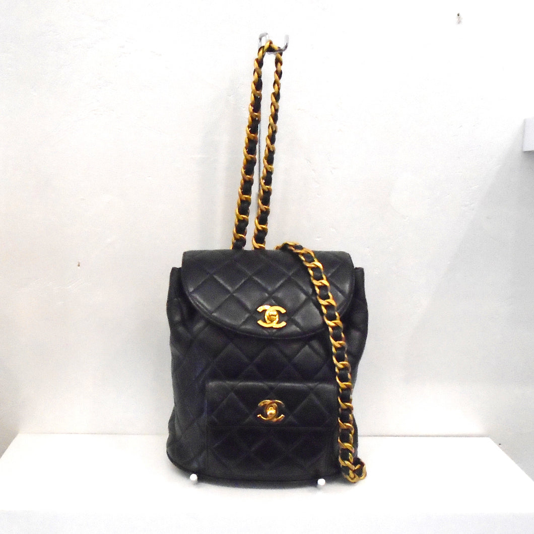 This Chanel Vintage Lambskin Backpack is Black with gold latches. It has a front pocket on the lower body of the bookbag. The straps are made of gold plated chain with a black leather strip woven through it.