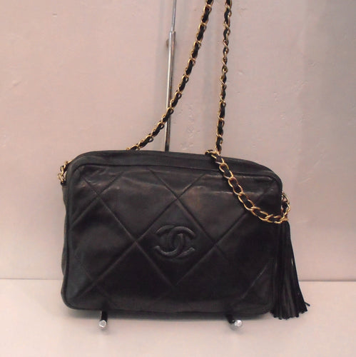 This Chanel Vintage Lambskin Camera Bag is a black bag that has a large diamond pattern with the CC logo in the center of the middle diamond. It has gold hardware and the shoulder chain has a black leather strip interwoven between the chain links. This bag also has a Tassle.