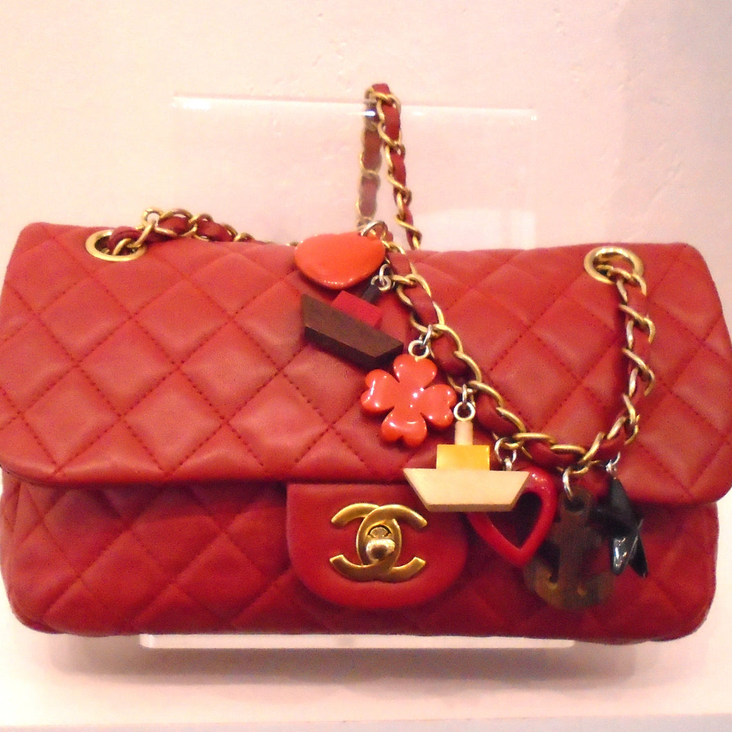 This Chanel Vintage Charm Lambskin Single Flap has a diamond pattern and a tab on middle of flap with CC logo. It has gold hardware with a red leather strip interwoven between the links and wooden charms of hearts, flowers, ships and anchors attached to the chain. 