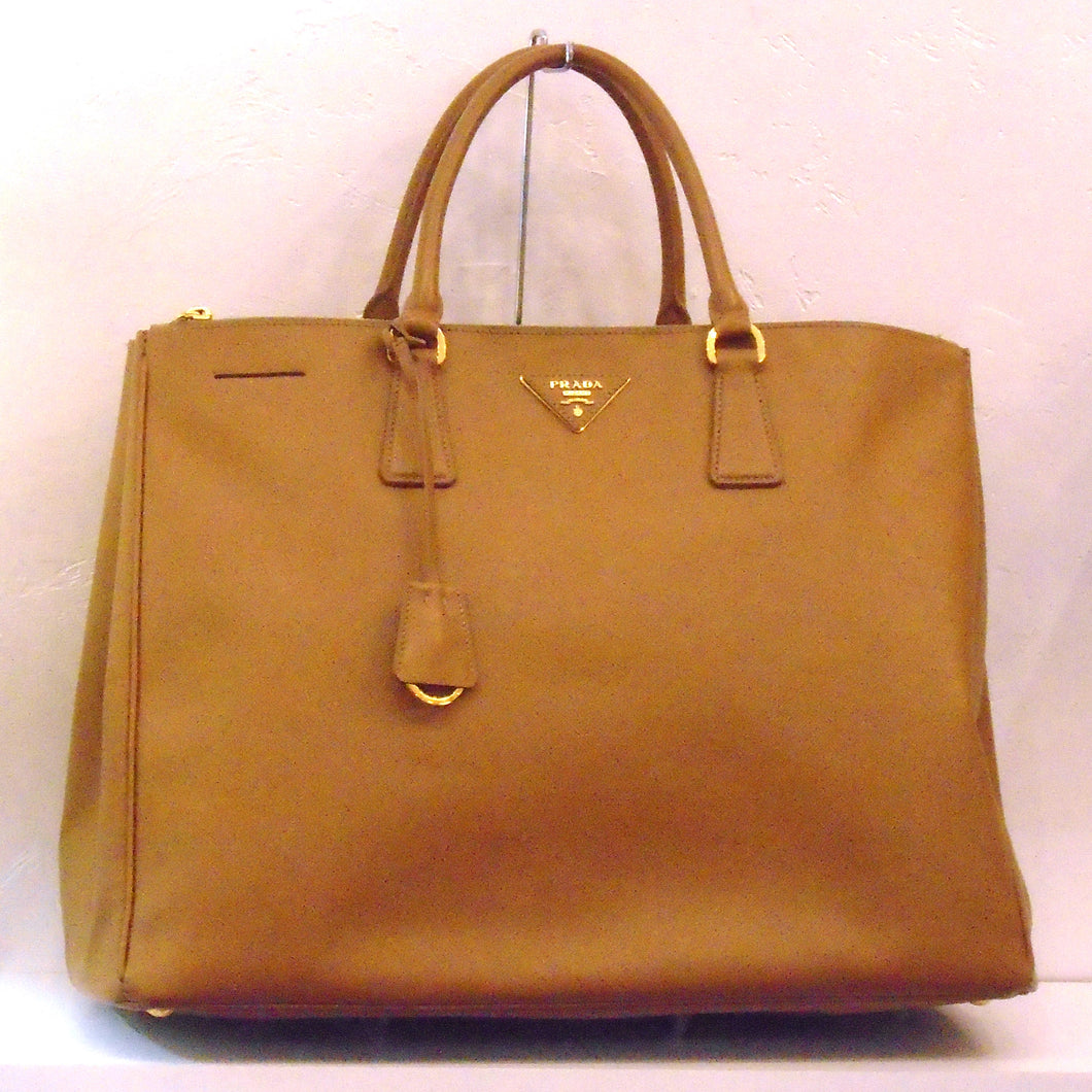 This Prada Caramel Saffiano Leather Large Tote has gold hardware, double carring handles and a key holder attached to the handles. It has a small guitar pick shaped prada name tag on the front and the inside is a silky green.